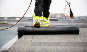 Pracownik z palnikiem montuje papę na dachu. Widoczna jest mineralna nawierzchnia i rolka materiału izolacyjnego.