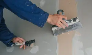 Ręce w niebieskim kombinezonie nakładają gładź wapienną na ścianę za pomocą szpachelek.