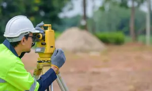 Geodeta w kasku i kamizelce odblaskowej pracuje z tachimetrem na budowie.