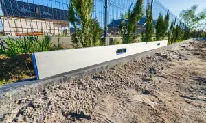 Poziomica na krawężniku podczas plantowania terenu. W tle ogrodzenie i dom w budowie.
