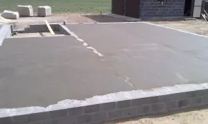 Świeżo wylany chudy beton na fundamencie domu. W tle widać materiały budowlane i fragment budynku.