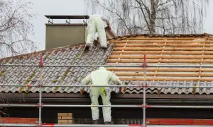Dwóch pracowników w kombinezonach usuwa stare pokrycie dachowe, prawdopodobnie eternit, z dachu domu.