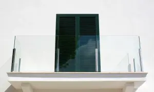 Nowoczesny balkon z przeszkloną balustradą i ciemnozielonymi okiennicami. Okapnik balkonu zapewnia ochronę przed deszczem.
