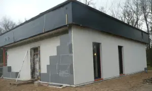 Budowa domu ze styropianu w trakcie. Widoczne białe bloczki, szare elementy fasady i czarna folia na dachu.