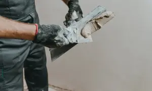 Ręce w rękawiczkach nakładają gładzie na ścianę za pomocą szpachli.