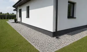 Biały dom z żwirową opaską wokół fundamentów i zielonym trawnikiem.