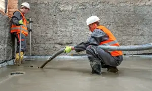 Dwóch budowlańców w kamizelkach odblaskowych wylewa pianobeton. Jeden z nich kieruje wąż, drugi kontroluje poziom.
