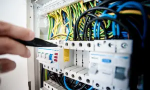 Ręka majstruje przy bezpiecznikach, tworząc instalację elektryczną w domu. Kolorowe przewody tworzą plątaninę.