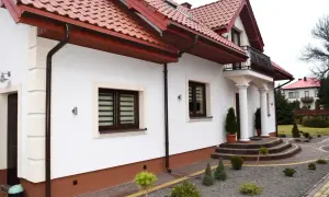 Elegancki dom z białym tynkiem i brązowym cokołem. Podjazd i schody z kostki brukowej, wokół zadbane krzewy. Idealny przykład, z czego zrobić cokół wokół domu.