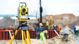 Żółty przyrząd geodezyjny na statywie na placu budowy, gdzie pracownicy w kaskach wykonują swoje zadania.