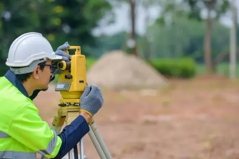 Geodeta w kasku i kamizelce odblaskowej pracuje z tachimetrem na budowie.