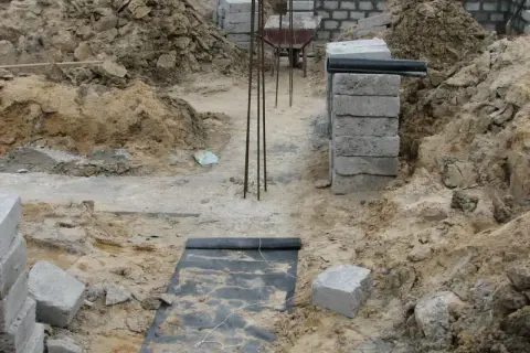 Budowa domu w toku. Widać wykopy, zbrojenie i materiały budowlane. Fundament czeka na zalanie.