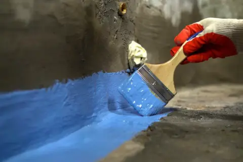 Rękawica w czerwonej rękawicy nakłada niebieski szlam hydroizolacyjny na betonową powierzchnię, tworząc barierę ochronną.
