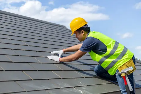 Dekarz w żółtym kasku i kamizelce odblaskowej układa dachówki. To właśnie dekarz dba o szczelność dachu.