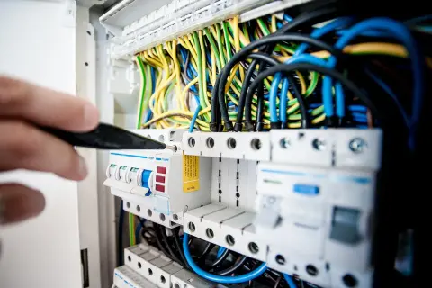Ręka majstruje przy bezpiecznikach, tworząc instalację elektryczną w domu. Kolorowe przewody tworzą plątaninę.