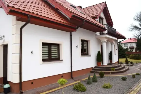 Elegancki dom z białym tynkiem i brązowym cokołem. Podjazd i schody z kostki brukowej, wokół zadbane krzewy. Idealny przykład, z czego zrobić cokół wokół domu.