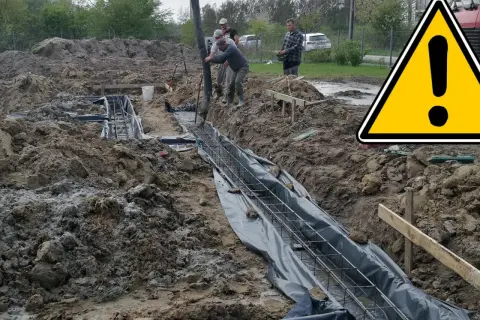 Pracownicy wylewają beton na zbrojenie, tworząc fundamenty domu. Teren budowy jest błotnisty.