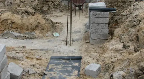 Budowa domu w toku. Widać wykopy, zbrojenie i materiały budowlane. Fundament czeka na zalanie.