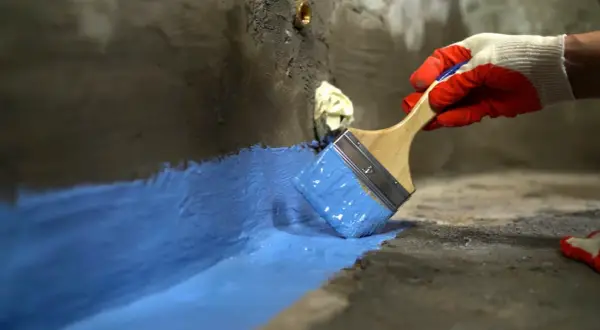 Rękawica w czerwonej rękawicy nakłada niebieski szlam hydroizolacyjny na betonową powierzchnię, tworząc barierę ochronną.
