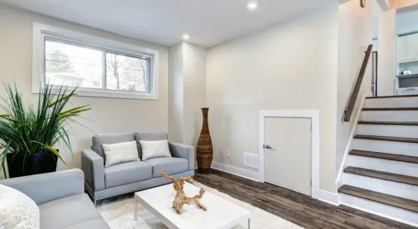 Przytulny suteren z szarymi sofami, białym stolikiem kawowym i dywanem. Obok schody prowadzące na górę.