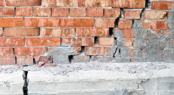 Uszkodzona ceglana ściana z pękniętym betonowym fundamentem.