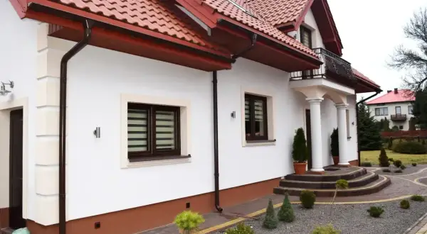 Elegancki dom z białym tynkiem i brązowym cokołem. Podjazd i schody z kostki brukowej, wokół zadbane krzewy. Idealny przykład, z czego zrobić cokół wokół domu.