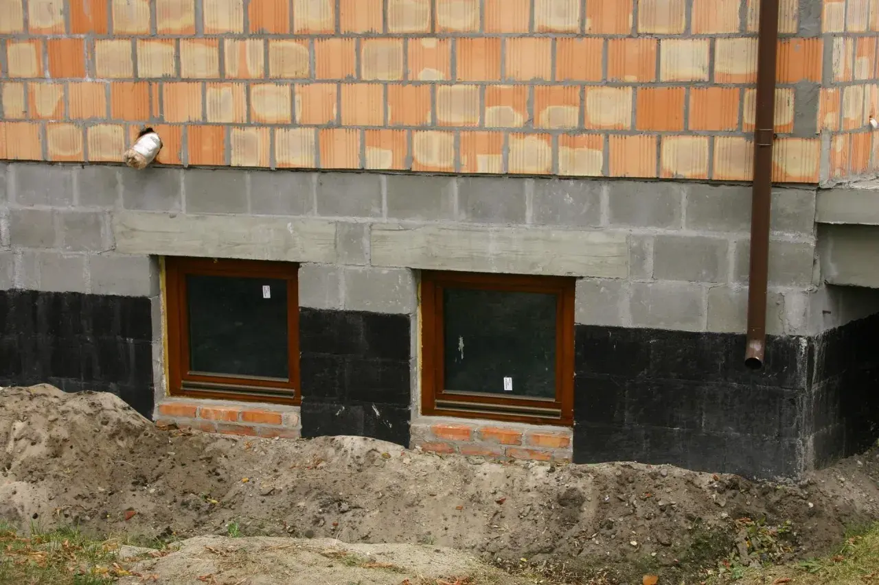Prace przy izolacji fundamentów w starym domu. Dwa okna piwniczne, czarna izolacja przeciwwilgociowa i nowe mury.
