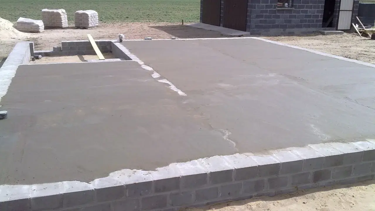 Świeżo wylany chudy beton na fundamencie domu. W tle widać materiały budowlane i fragment budynku.