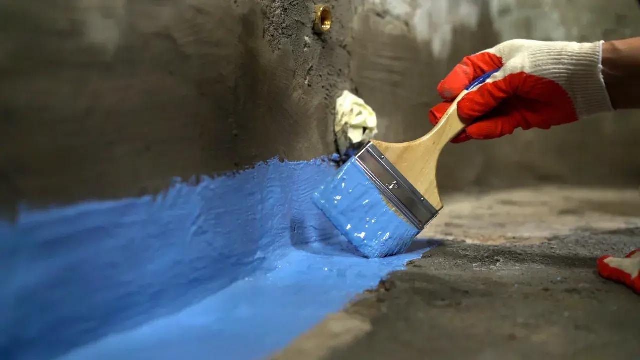 Rękawica w czerwonej rękawicy nakłada niebieski szlam hydroizolacyjny na betonową powierzchnię, tworząc barierę ochronną.