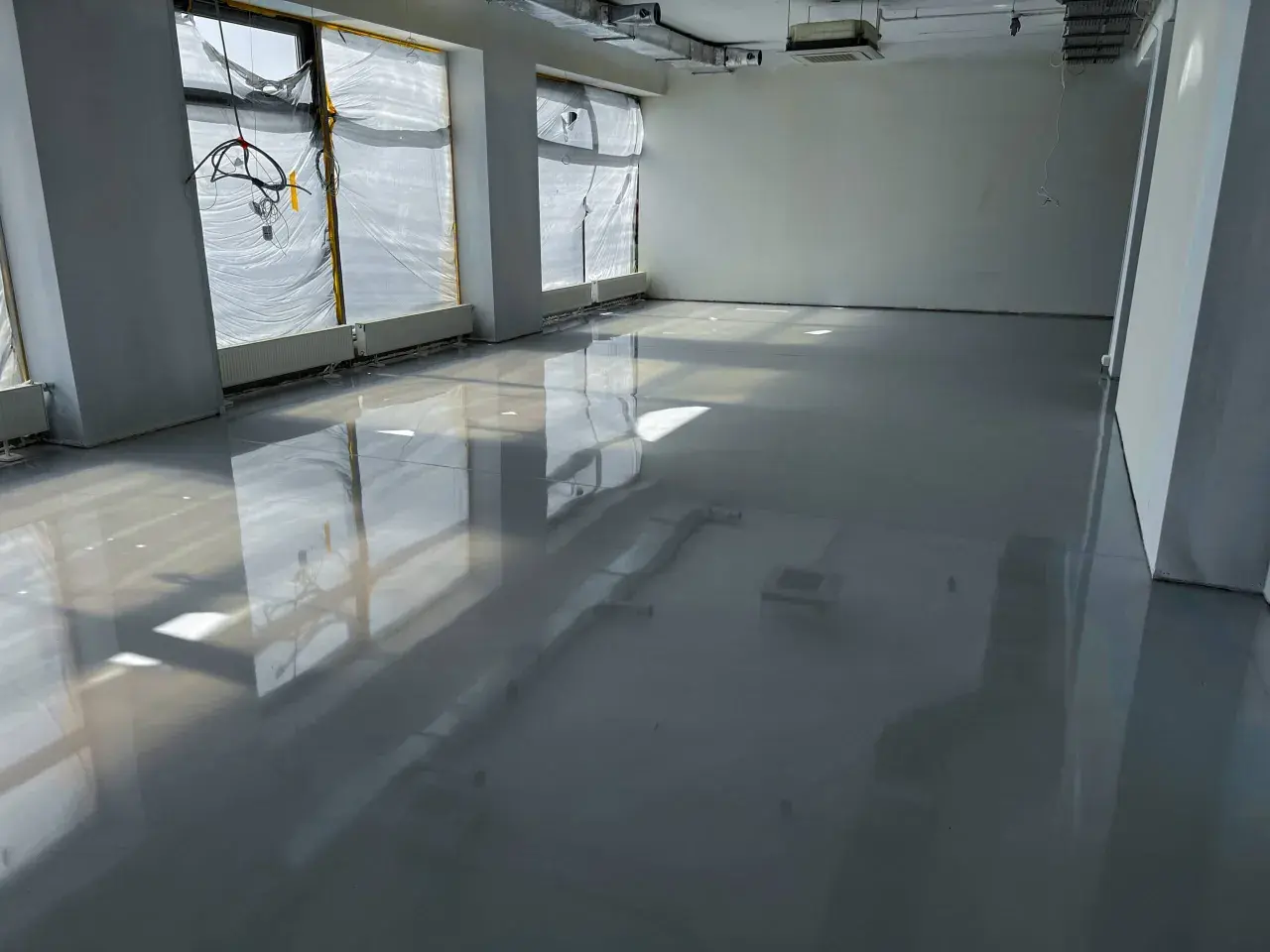 Lśniąca podłoga z żywicy w pomieszczeniu w budowie. Widać odbicia okien i instalacji.