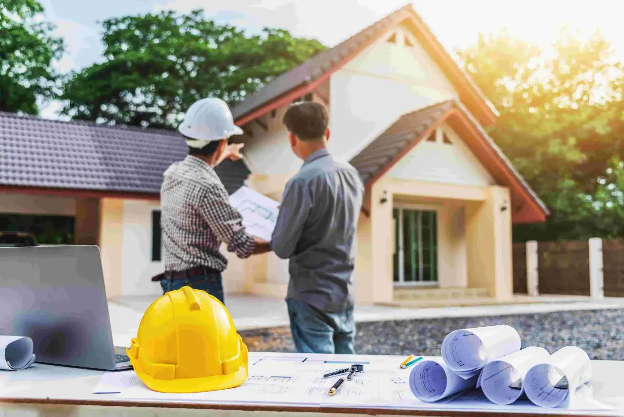 Inspektor nadzoru inwestorskiego omawia plany budowy domu z pracownikiem. Na stole leżą plany, kask i laptop.