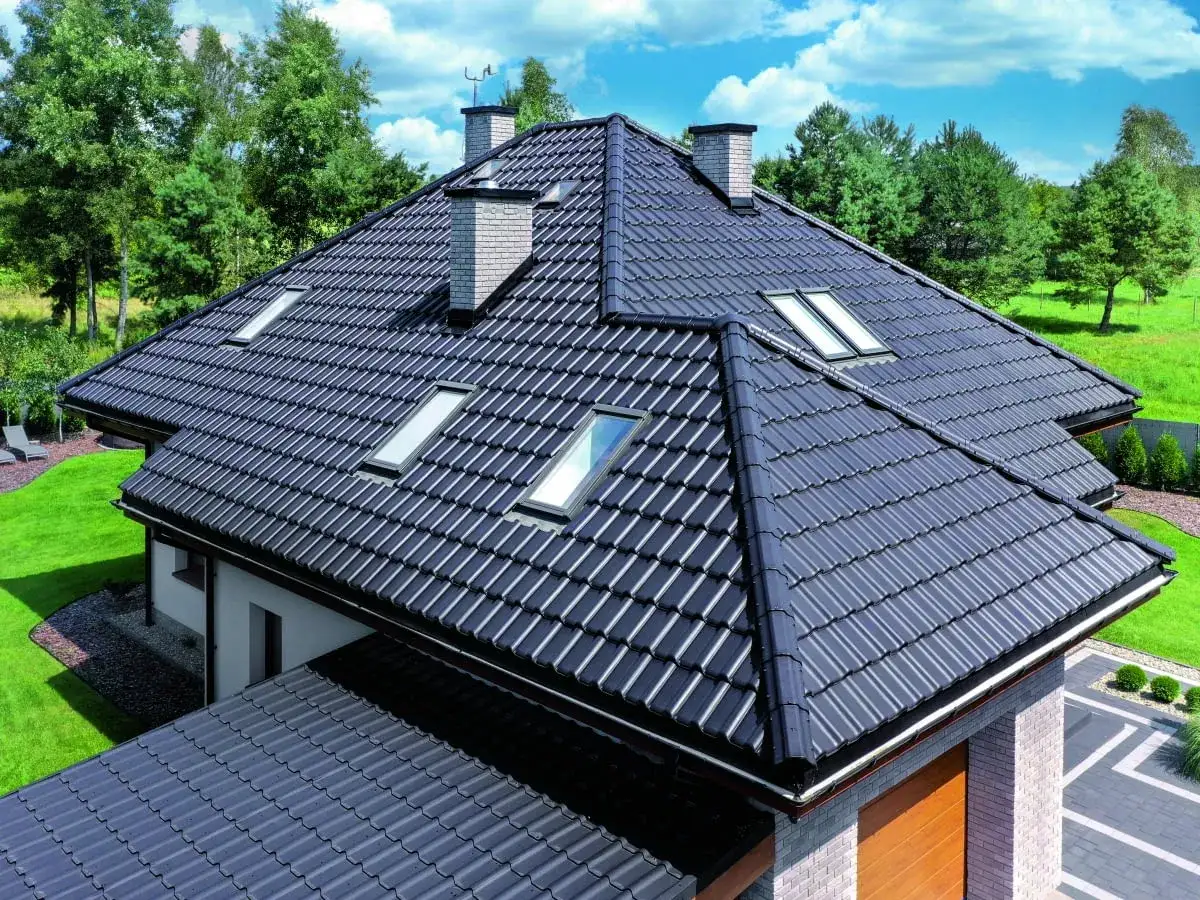 Nowoczesny dach wielospadowy z ciemnej dachówki, z kilkoma oknami połaciowymi i dwoma kominami, otoczony zielonym ogrodem.