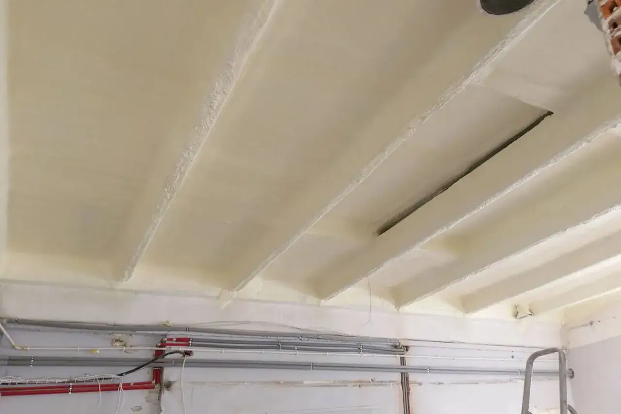 Ocieplenie stropu betonowego pianką PUR. Widoczne belki stropowe pokryte izolacją, pod nimi instalacje.