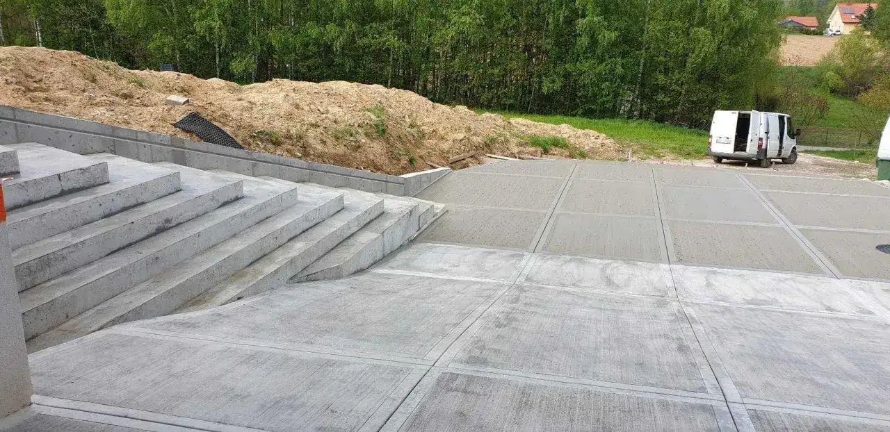 Schody i plac wykonane z betonu szczotkowanego, otoczone zielenią i skarpą z piasku. W tle biały van.