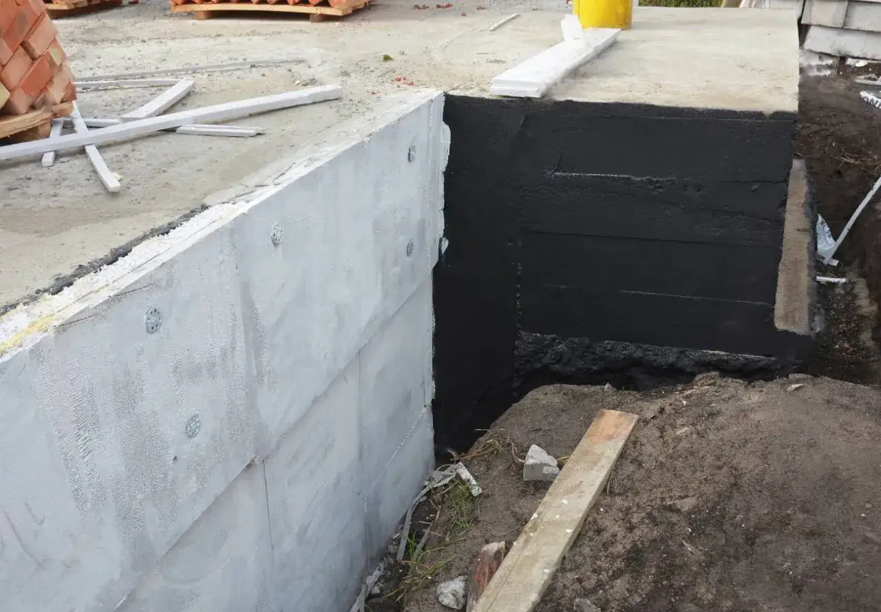 Fundament budowlany z płyt betonowych, jedna strona zagruntowana na czarno, druga biała. W tle materiały budowlane, w tym masa kmb.