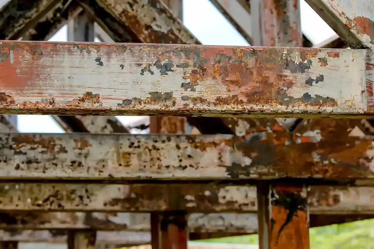 Metalowe belki konstrukcji z widoczną łuszczącą się farbą i rdzą. Widać ślady postępującej korozji, która niszczy materiał.