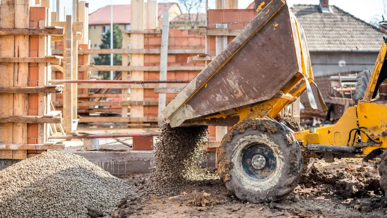 Żółta ładowarka zrzuca żwir na placu budowy. W tle widać drewniane szalunki i ceglane ściany, elementy budowy, która powstaje z materiałów Lafarge.