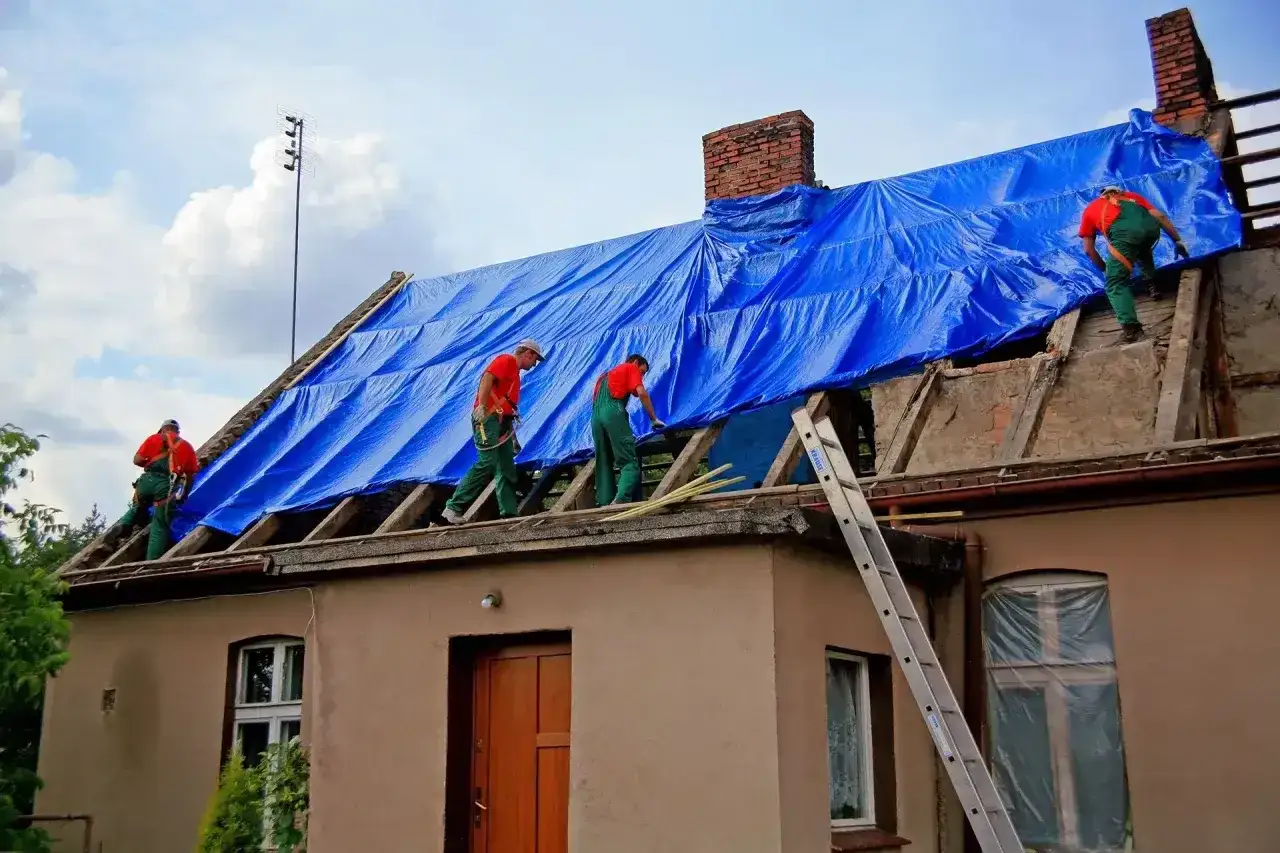 Pracownicy zabezpieczają dach niebieską plandeką. Remont dachu wymaga pozwolenia na budowę? Sprawdź przepisy.