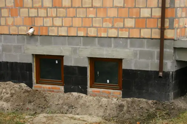 Prace przy izolacji fundamentów w starym domu. Dwa okna piwniczne, czarna izolacja przeciwwilgociowa i nowe mury.