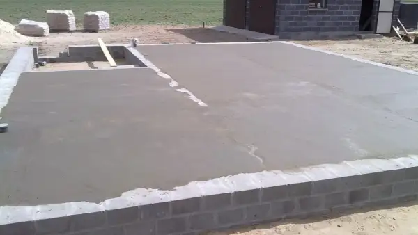 Świeżo wylany chudy beton na fundamencie domu. W tle widać materiały budowlane i fragment budynku.
