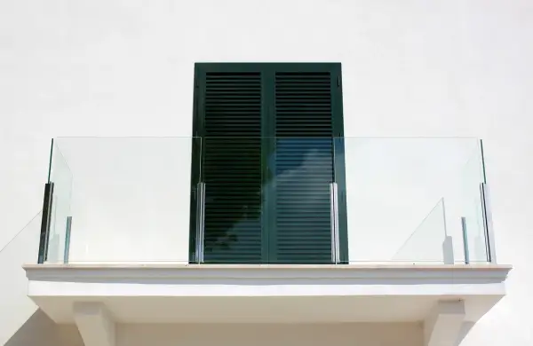 Nowoczesny balkon z przeszkloną balustradą i ciemnozielonymi okiennicami. Okapnik balkonu zapewnia ochronę przed deszczem.