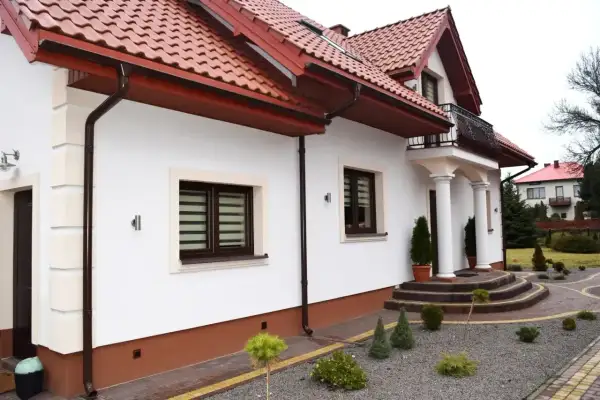 Elegancki dom z białym tynkiem i brązowym cokołem. Podjazd i schody z kostki brukowej, wokół zadbane krzewy. Idealny przykład, z czego zrobić cokół wokół domu.